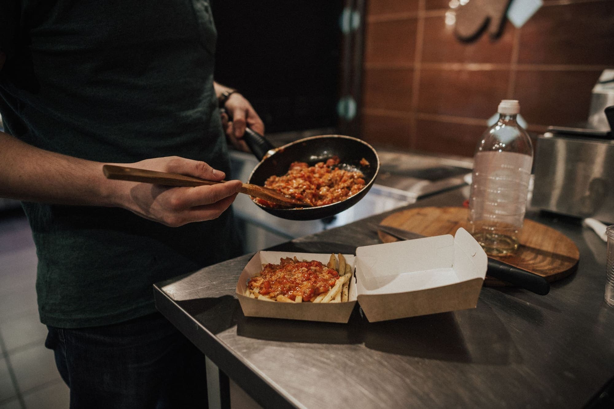 A cook spooning sauce over chips in a takeaway box on a steel kitchen counter